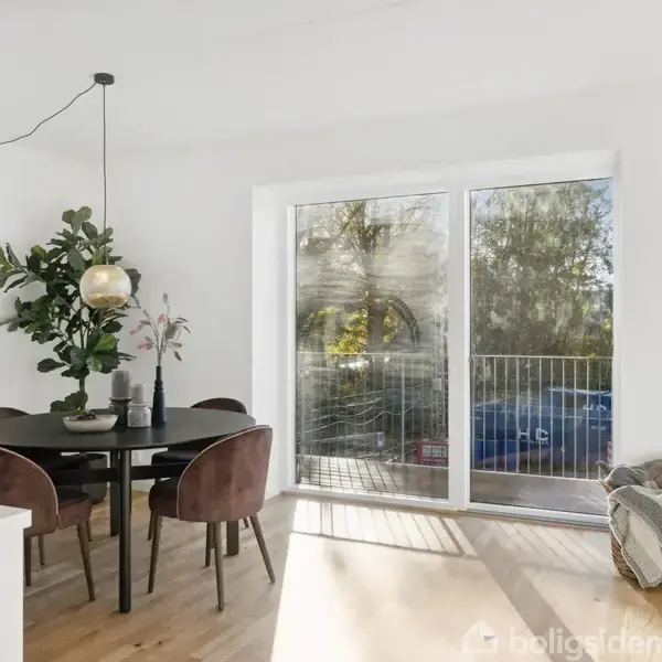 Et rundt bord dekoreret med en lampe og planter står i et lyst rum foran store glasdøre, der fører ud til en balkon med udsigt til træer.