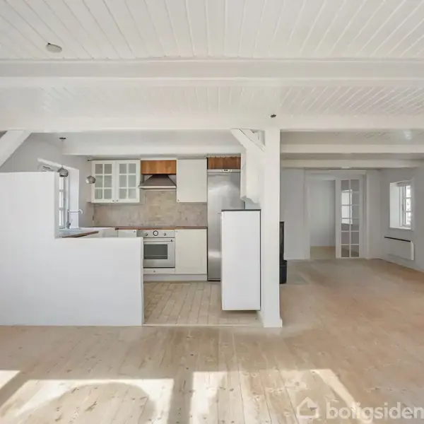 Et lyst køkken med åbne hylder og hvide skabe ligger i en rummelig stue med trægulv. Store vinduer lader naturligt lys strømme ind, og rummet virker indbydende.