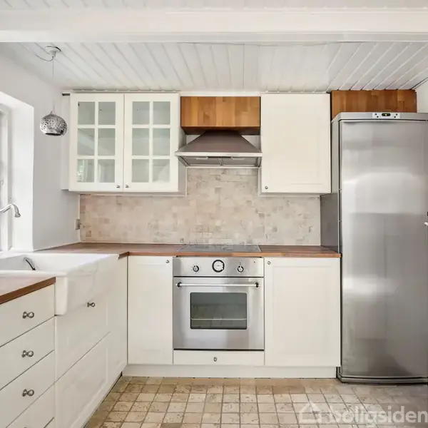 Hvidt køkken med stålkomfur og ovn indbygget i træbordplade, køleskab ved siden af, overskabe med glaslåger. Lysindfald fra vindue til venstre, flisegulv i beige toner.