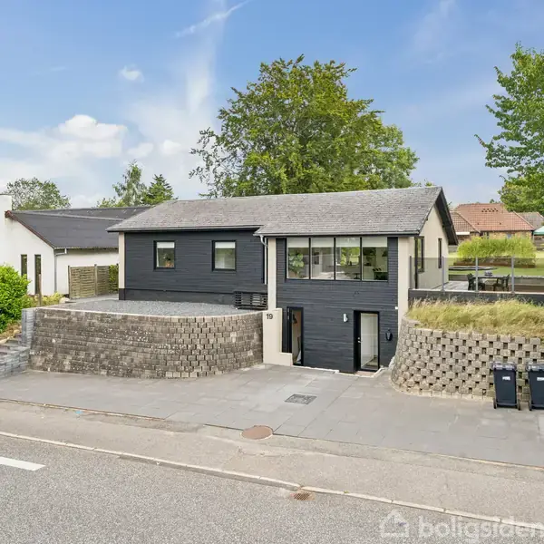 Et moderne, sort træhus med store vinduer står på en hævet grund omkranset af stensætninger og grønne træer langs en stille vej.