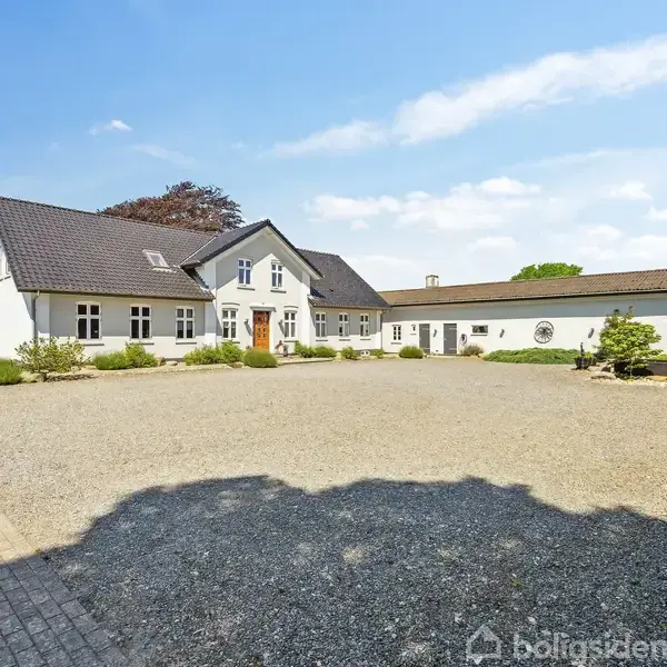 En hvid bygning med sort tag står på en grusplads, omkranset af grønne buske og træer. Bygningen har flere vinduer og en centralt placeret hoveddør.