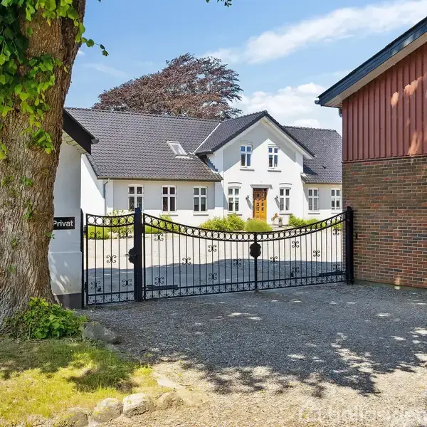 Et hvidt hus med mørkt tag er set gennem en sort metalport. Til venstre står et stort træ ved siden af et skilt med teksten "Privat". Grusvej og grønne omgivelser.