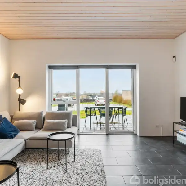 En grå sofa står i en stue med moderne indretning. En lampe lyser hjørnet op ved sofaen. Et glasdørparti fører ud til en terrasse med havemøbler.