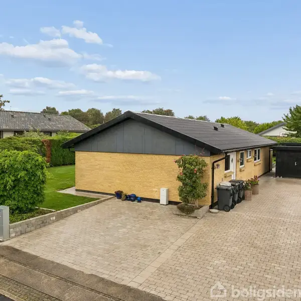 Et gult murstenshus står stille i et villakvarter. Foran huset er en stor flisebelagt indkørsel med affaldscontainere og en sort carport. Grønt buskads omkranser ejendommen.