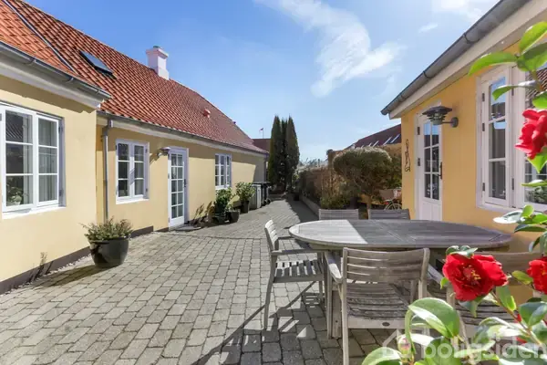 Rundt havebord med stole står på en brostensbelagt terrasse mellem to gulmalede huse med røde tegltage. Forskellige planter omkranser området, og himlen er klar med få skyer.