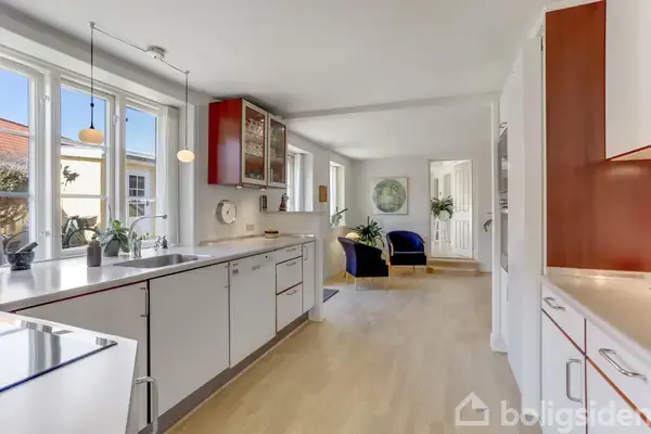 Det lyse køkken har hvide skabe, en række vinduer med planter, og hængende lamper. Et område med blå sofaer og planter ses i baggrunden.