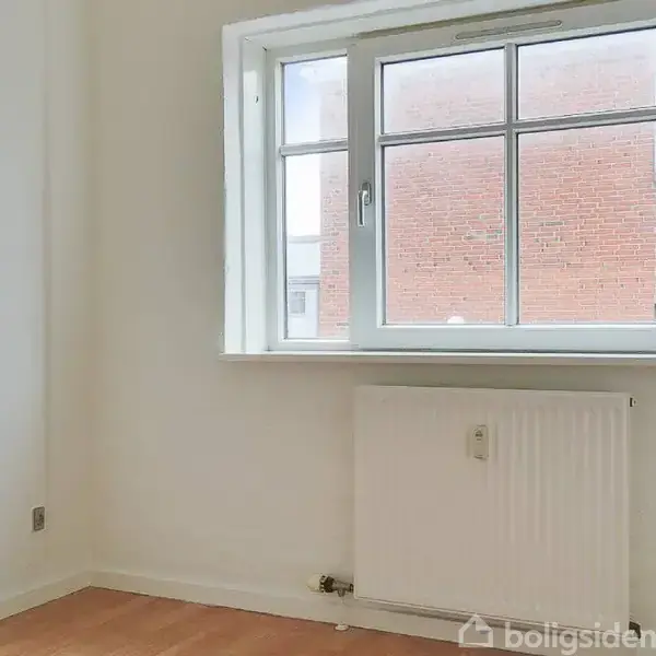 Vindue står åbent udad mod en murstensbygning. Under vinduet er der en hvid radiator. Vægge og gulv er lyse i farvetonen. Ingen tekst er til stede.