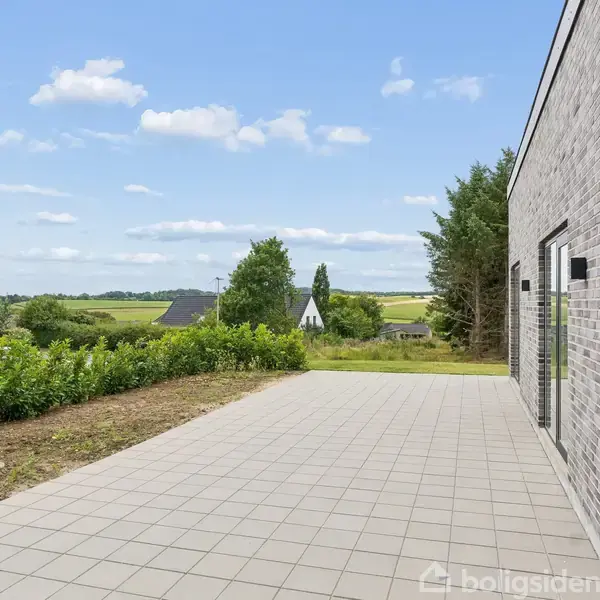 En terrasse med grå fliser støder op til en moderne murstensbygning. I baggrunden er der grønt landskab med træer og marker under en let skyet himmel.