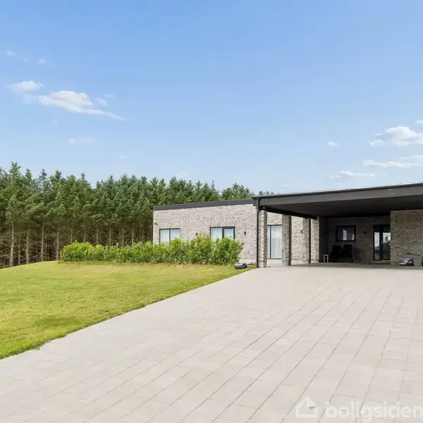 En moderne bungalow står stille i et åbent landskab med en lang indkørsel og en velholdt græsplæne. Omkring huset er der en skovagtig baggrund.