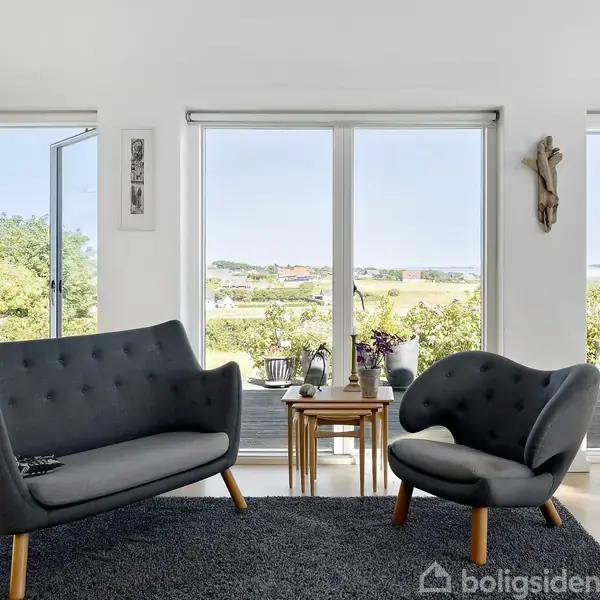 To sofagruppe med grå polstring står på et mørkt tæppe i en lys stue med store vinduer, der åbner op til en terrasse med udsigt til grønne omgivelser.