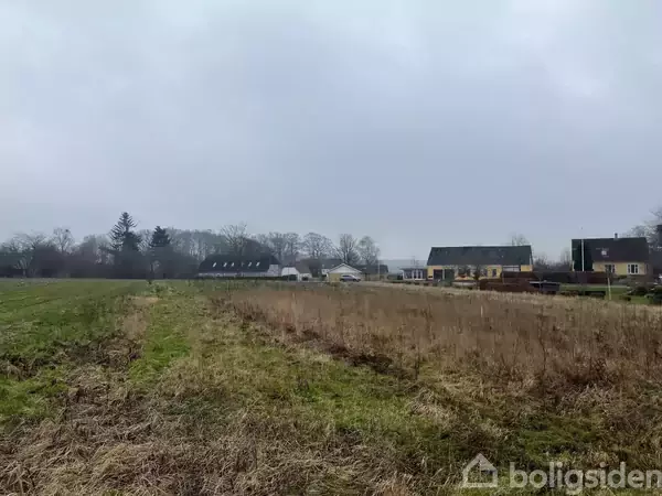 En flad mark ligger ubenyttet med tørt græs, mens huse ses i baggrunden. Himlen er overskyet, og der er træer langs horisonten, hvilket skaber en landlig atmosfære.