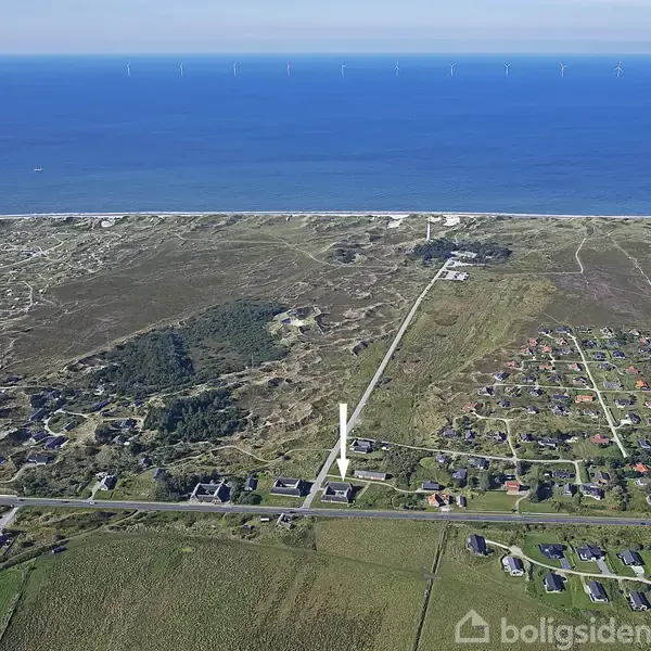 Kystlandskab med huse spredt ud over marker og skove, hvor de ligger i nærheden af en strandlinje. Havet ses i baggrunden med en række havvindmøller i horisonten.
