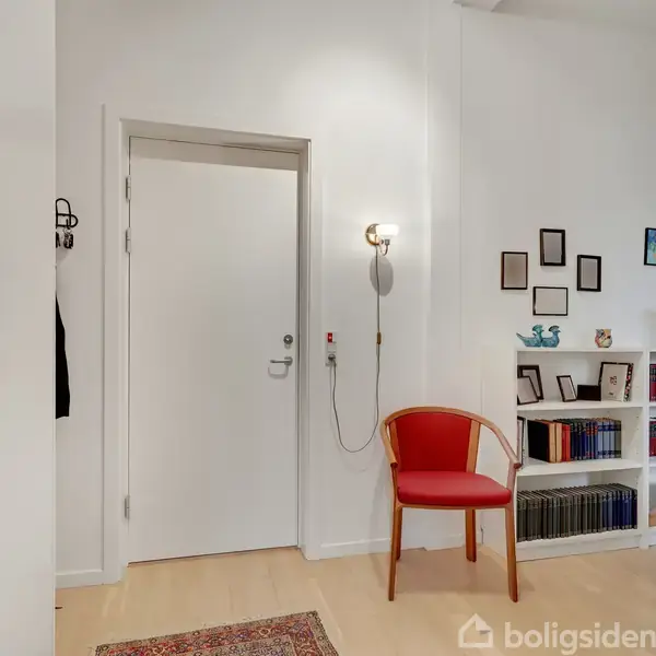 Dør står lukket i hvid væg ved siden af rød stol, oplyst af væglampe. Hylde med bøger og pynt på modsatte væg, rammer hænger over den. Tæppe på trægulv.