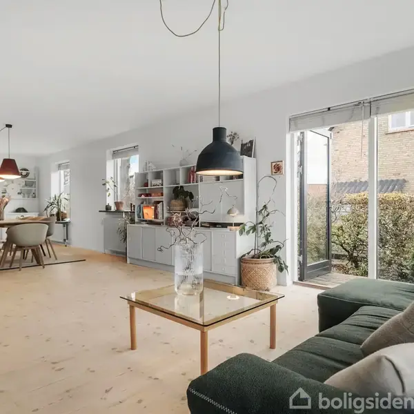 En moderne stue med en grøn sofa og glasbord under en sort pendellampe. Store vinduer giver udsigt til en have. I baggrunden er et spisebord med stole.