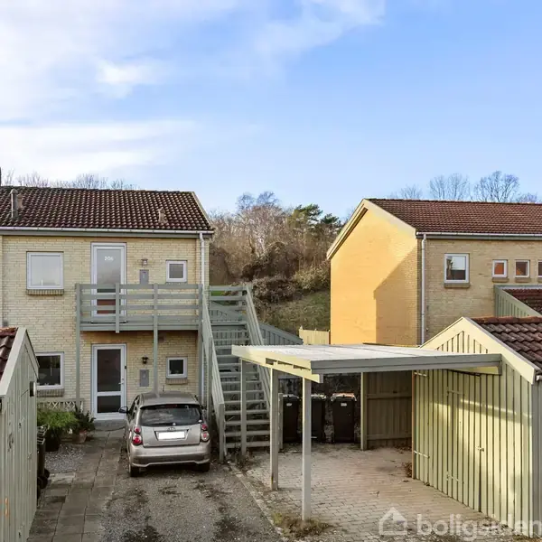 To biler holder parkeret ved siden af hinanden foran et beboelseskompleks med lys murstensfacade og trapper. I baggrunden ses en grøn beplantning og delvist overskyet himmel.