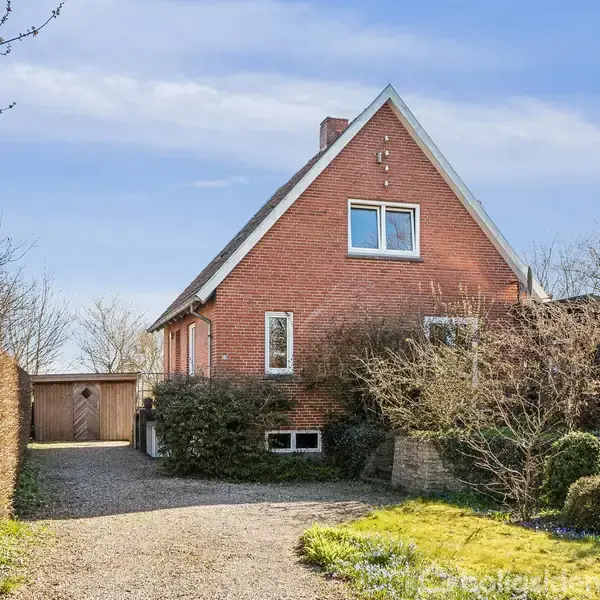 Rødt murstenshus med gavltag står stille på en grusvej mellem buske og træer i et villakvarter. Huset har flere vinduer og en tilhørende enkelt garage i baggrunden.