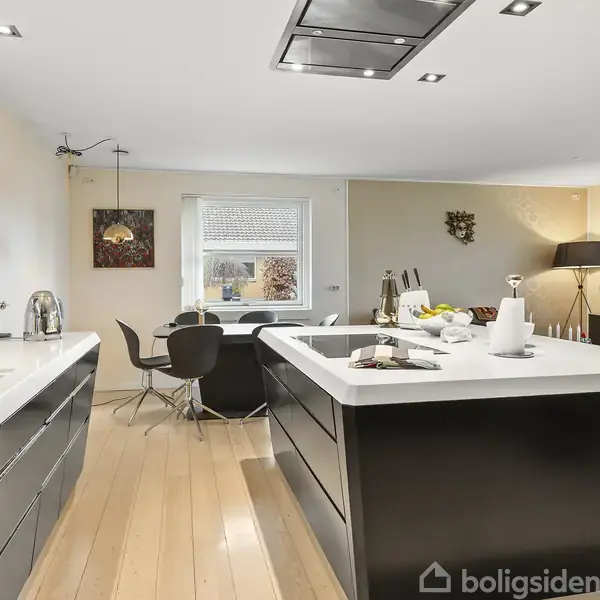 Et moderne køkken med sort køkkenø og indbygget komfur; lyse træplanker på gulvet; spisebord med sorte stole ved vinduet; dekorativ lampe og kunst på væggen.