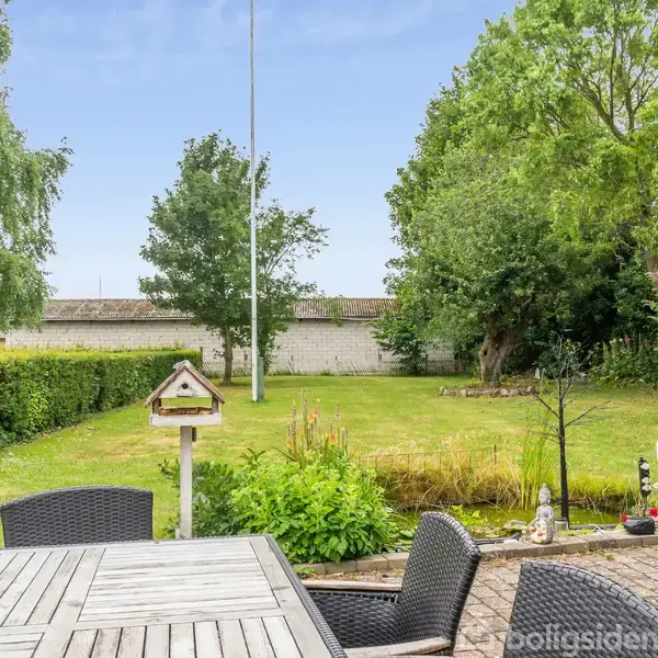 Udendørs terrassebord med sorte stole står på en flisebelagt terrasse. En velplejet have omgivet af buske og træer samt et fuglehus findes i baggrunden.