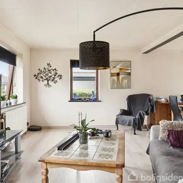 Stue med et gråt sofaarrangement og sofabord i træ, en fladskærm på et mediebord. Mørklægningsgardiner dækker to vinduer. Spisebord og stole i baggrunden ved en lysekrone.