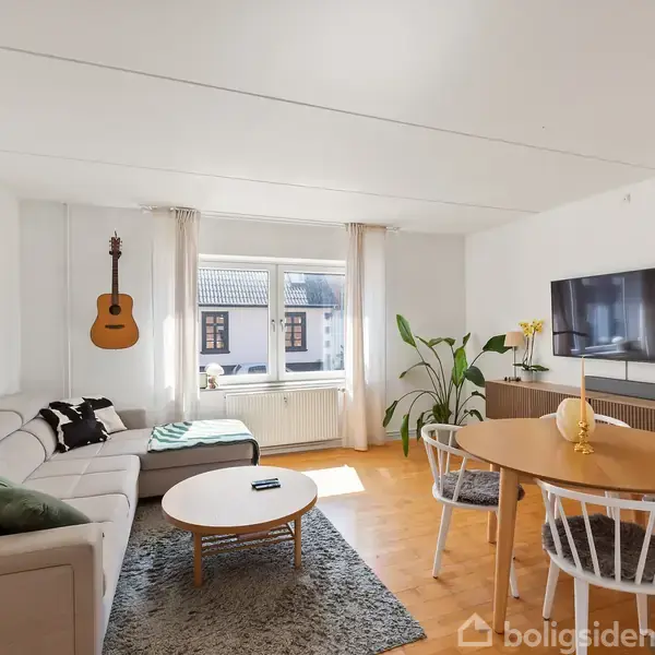 En lys stue med en L-formet sofa, et rundt sofabord og en vægmonteret guitar. Et tv hænger på væggen over et skab, og der er et spisebord med stole.