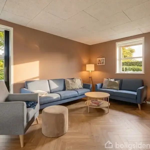 Stue med to blå sofaer, en grå lænestol og en rund træbord. Lamper lyser rummet. Vindue viser grønt have. Varm belysning skaber hyggelig atmosfære.