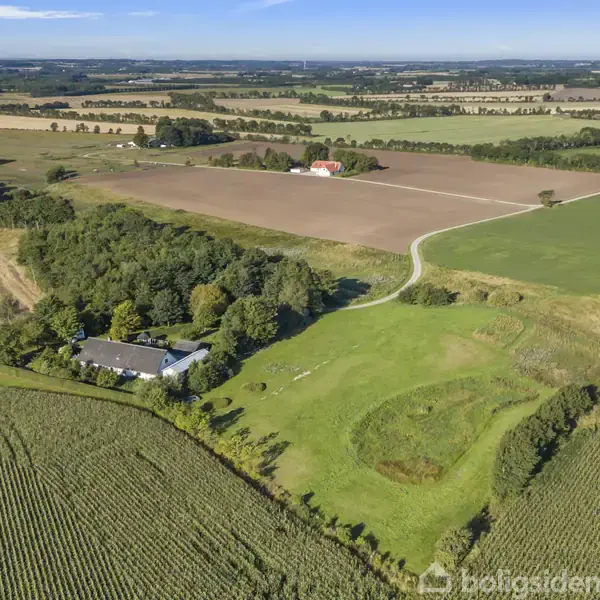 Et hvidt hus ligger omkranset af træer og marker. Veje snor sig gennem landskaberne, som er præget af opdyrkede marker og skove i det fjerne under en blå himmel.
