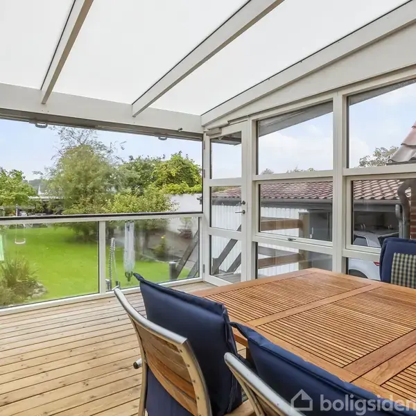 En overdækket terrasse med træbord og blå stole står i forgrunden. Den hvide overdækning og store glaspartier giver udsigt til en grøn have med træer i baggrunden.