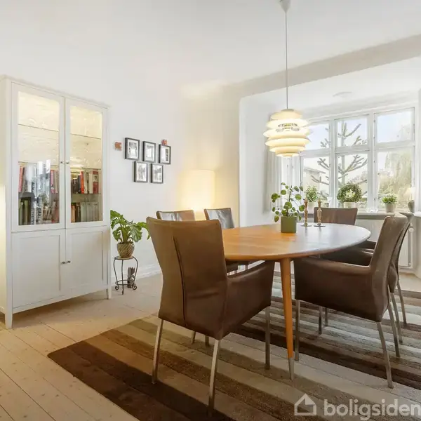 Et rundt spisebord med fem stole står på et tæppe i en lys spisestue. Der er planter i vinduet og en lampe hænger over bordet.