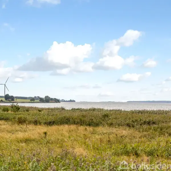 Vingemølle står stille på en mark med højt græs. I baggrunden ses en stor sø under en blå himmel med spredte skyer.