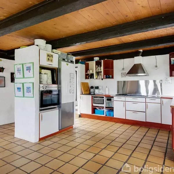 Et køkken med hvide skabe og rustfri ståloverflade er udstyret med ovn og emhætte. Fliser dækker gulvet, og rummet har synlige træbjælker i loftet.