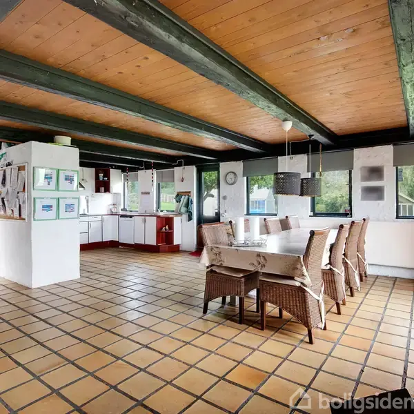 Et spisebord med kurvestole står i et rummeligt køkken-alrum med fliser og træbjælker. Store vinduer giver udsigt til træer udenfor, mens indrammede billeder hænger på væggene.