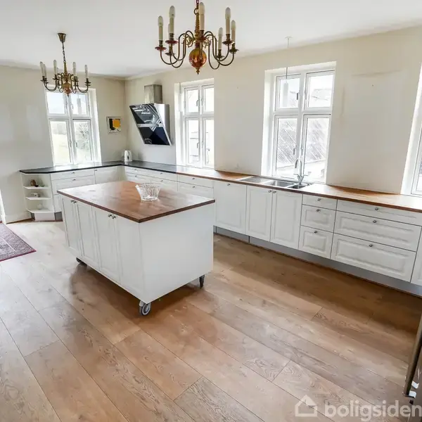 Rødt murstenshus med hvide vinduer står på en velplejet græsplæne. Det er omgivet af grønne træer under en klar blå himmel med hvide skyer.