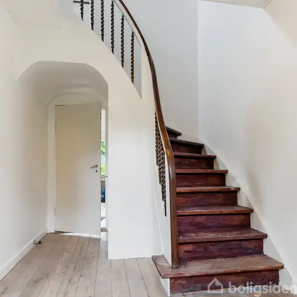 Trætrappe leder opad i en hvidmalet gang. En åben dør til venstre viser et andet rum. Mørkt gelænder følger trappens kurve. En delvis udsigt af et vindue kan ses.