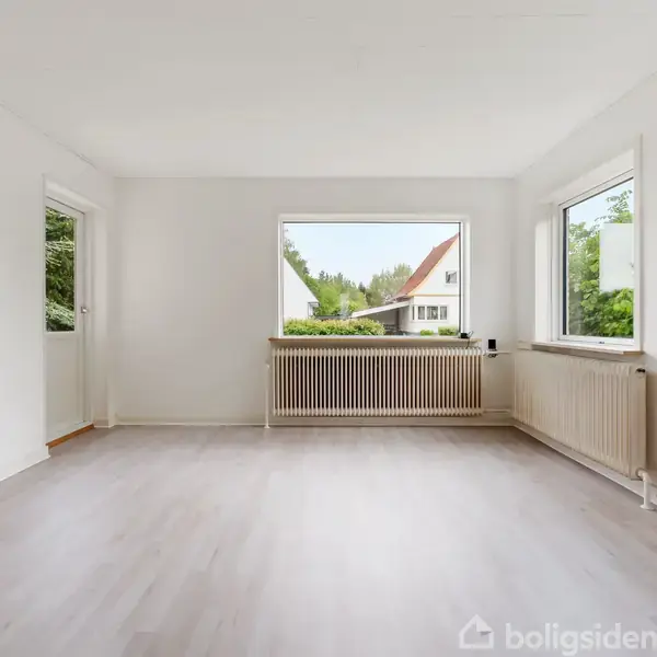 Et tomt, lyst værelse med trægulv, store vinduer med udsigt til en grøn omgivelser, og radiatorer under vinduerne. Der er en åben dør til venstre. Ingen tekst til stede.