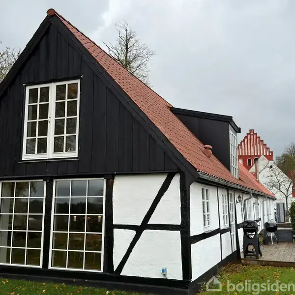 Sort-hvid bindingsværkshus med rødt tegltag står stille i en have med træer og plæne. En terrasse med havemøbler er placeret ved siden af huset.