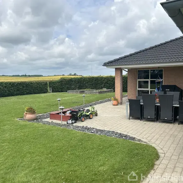Terrasse med flisebelægning og havemøbler ved et rødt murstenshus. Legetøjstraktor på græsplæne. Bagved hækken ses åbne marker. Himlen er overskyet.