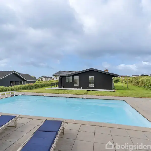 Svømmebassin med blå solsenge omkring står i forgrunden. I baggrunden ses et sort træhus omgivet af grønne plæner og hække, under en overskyet himmel.