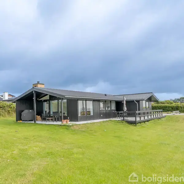 Et sort sommerhus står på en grøn græsplæne under en overskyet himmel. Huset har store vinduer og en veranda, omgivet af tætte buske og græsområder.