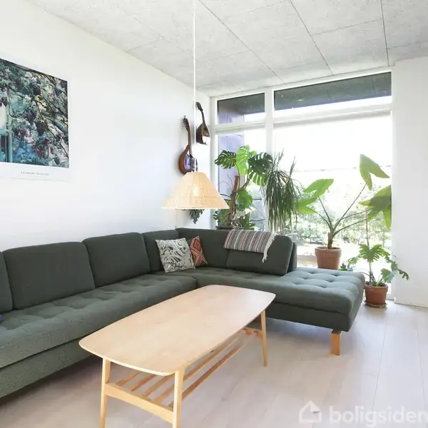 En grøn sofa står stille i et lyst rum med store vinduer og mange grønne planter. En træbordplade hviler foran sofaen, mens en hængende lampe og kunst på væggen skaber stemning.