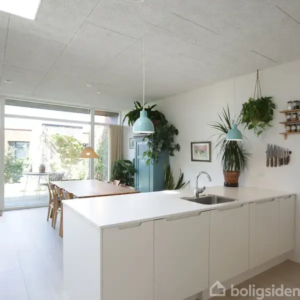 Køkken med hvide skabe og håndvask foran. Spisesektion med bord og stole ved vindue mod haven. Planter hænger fra loftet, hylder med krydderiopbevaring på væggen.