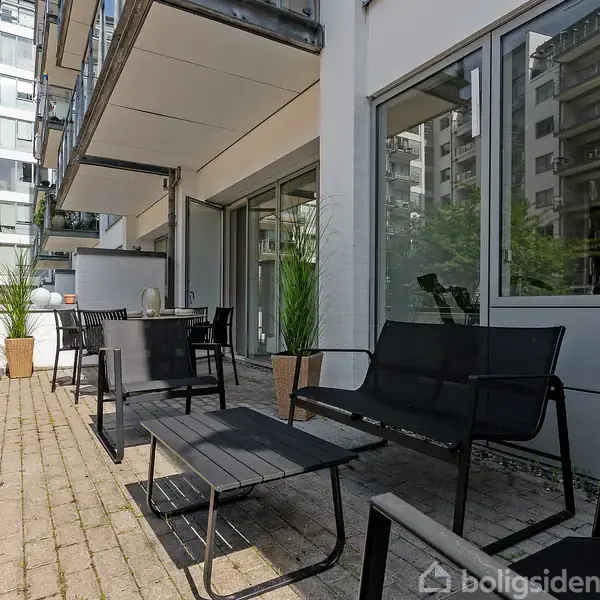 Sort terrassemøbel sæt på flisebelagt altan med to sorte stole og et bord. Moderne lejlighedskompleks i baggrunden. Store vinduer på bygningsfacaden reflekterer sollys.