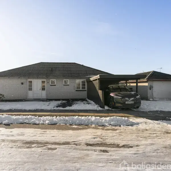En bil holder parkeret i en carport ved siden af et hvidt, enkeltliggende hus med sne omkring og en klar blå himmel i baggrunden.