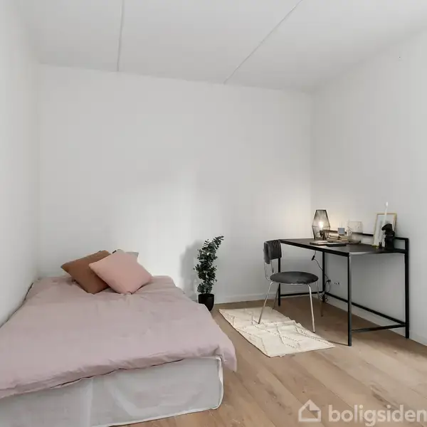 En enkeltseng med lyserødt sengetøj står i et lyst værelse. Ved siden af er der et sort skrivebord med en stol og lampe. En plante står ved siden af sengen.