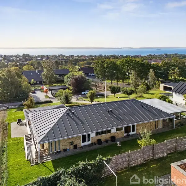 Hus med fladt tag og stor terrasse ses fra luften. Beliggende i et grønt kvarter med udsigt over vandet i baggrunden og omgivet af andet boligbyggeri og træer.