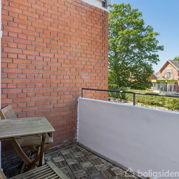 En lille altan med et bord og to stole står stille omgivet af røde mursten og glasvægge. Udenfor ses træer og et hus i baggrunden.