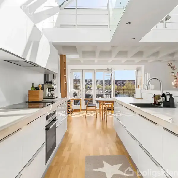 Et lyst køkken med hvide skabe og moderne apparater, herunder en ovn og kogeplade. Der er en glasvæg mod en balkon med udsigt til et byområde, og et spiseområde med træbord og stole.