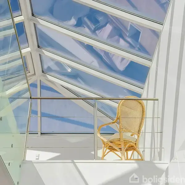 En kurvestol står stille på en moderne balkon under et glasloft, der lader dagslys trænge ind. Rummet er lyst og rummeligt med en minimalistisk æstetik.