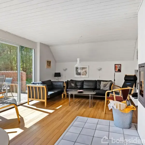 Stue med sort sofa og stole arrangeret omkring et sofabord. Store vinduer til venstre åbner mod en terrasse. Rummet har lyse trævægge og -gulv og et moderne interiør.