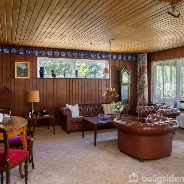 Stue med brune lædersofaer og et rundt træbord. En kamin og en standerlampe. Vægge dekoreret med tallerkner og billeder. Store vinduer giver udsigt til en grøn have.