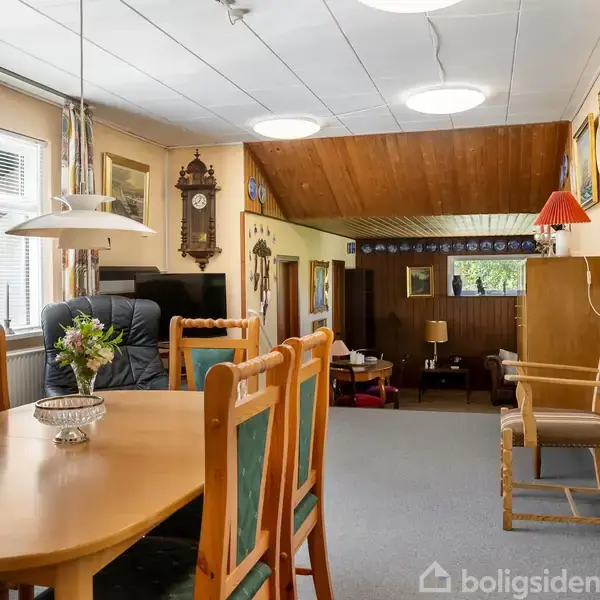 Spisebord med træstole står i en stue, hvor et ur hænger på væggen. Omkring er en sofa, stole og malerier. Vinduet til venstre lukker sollys ind.
